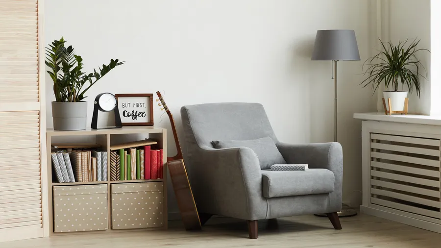 A cozy reading nook with gray armchair, wooden shelf with books, guitar, and a floor lamp.